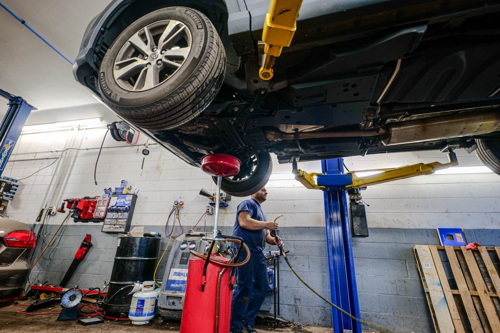 Mechanic working underneath a lifted car in an auto repair shop with various tools and equipment around.